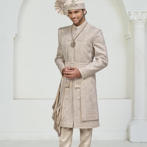 May include: A beige embroidered sherwani suit with matching pants and a turban. The long coat features intricate patterns and a draped sash. A gold-colored necklace adds a touch of elegance to the ensemble. The outfit is suitable for special occasions.