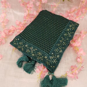 Könnte beinhalten: Gefaltetes, smaragdgrünes Textil mit Paillettenmuster und einem Samtrand mit goldfarbener Blumenstickerei. Das Textil hat passende grüne Quasten an den Ecken und wird auf einer weißen Oberfläche mit rosa floralen Akzenten präsentiert.