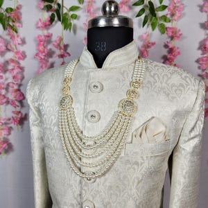May include: A cream-colored, patterned jacket with a matching pearl necklace. The necklace has multiple strands and gold-colored decorative elements. A white pocket square is in the jacket pocket. The mannequin has a size 38 label.