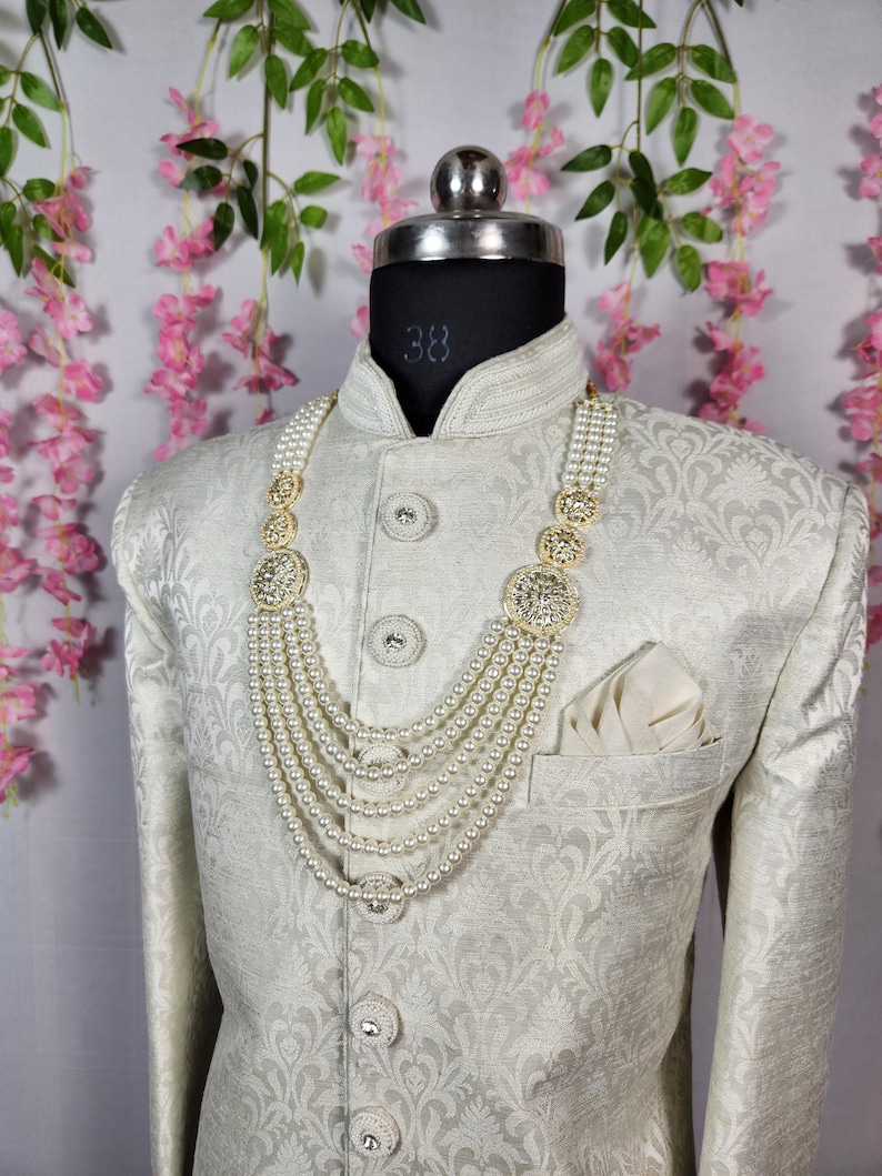 May include: A cream-colored, patterned jacket with a matching pearl necklace. The necklace has multiple strands and gold-colored decorative elements. A white pocket square is in the jacket pocket. The mannequin has a size 38 label.