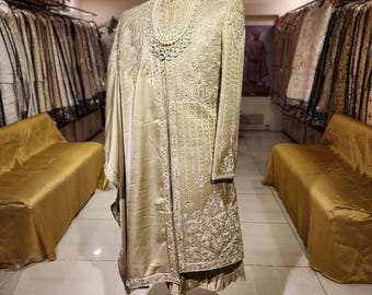Puede incluir: Un traje de boda bordado de color dorado, exhibido en un maniquí. El traje incluye una chaqueta larga, pantalones, un chal y un turbante con una pluma. Un collar de perlas adorna el cuello del maniquí. El traje se exhibe en una tienda de ropa.