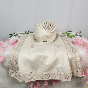 Könnte beinhalten: Ein elfenbeinfarbener Turban mit einem fächerförmigen Design und einem passenden Schal. Der Schal hat aufwendig bestickte Ränder und florale Muster. Die Artikel werden auf einer weißen Oberfläche präsentiert, die mit rosa Blumen geschmückt ist.