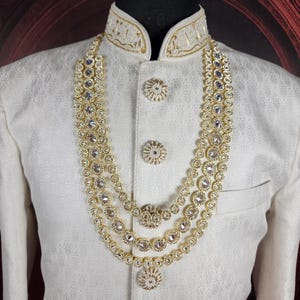 K&ouml;nnte beinhalten: Cremefarbene Jacke mit goldener Stickerei und dreireihiger Halskette. Die Halskette hat gro&szlig;e, klare Edelsteine in Gold gefasst. Die Jacke hat dekorative Kn&ouml;pfe und einen hohen Kragen mit goldenen Details.