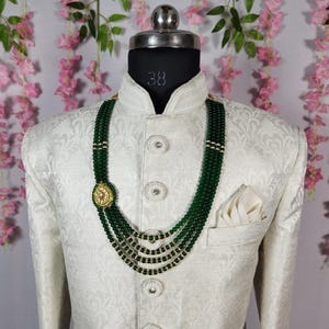 Puede incluir: Chaqueta color crema con un sutil estampado floral, combinada con un collar de cuentas verdes de varias hebras. El collar presenta detalles dorados y un colgante decorativo. Un pa&ntilde;uelo blanco est&aacute; visible en el bolsillo de la chaqueta.