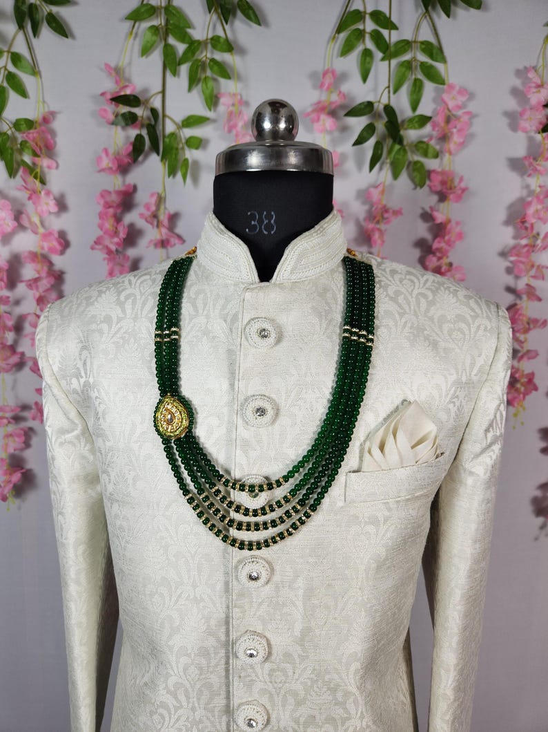 Puede incluir: Chaqueta color crema con un sutil estampado floral, combinada con un collar de cuentas verdes de varias hebras. El collar presenta detalles dorados y un colgante decorativo. Un pa&ntilde;uelo blanco est&aacute; visible en el bolsillo de la chaqueta.