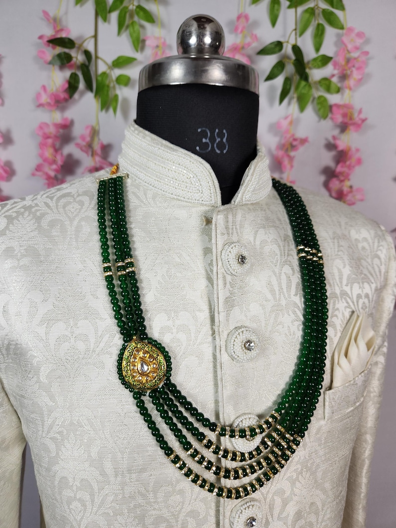 Puede incluir: Un collar de cuentas verdes con un colgante dorado con dise&ntilde;o de l&aacute;grima y detalles de piedras blancas. El collar se exhibe en un maniqu&iacute; que lleva una chaqueta blanca estampada.