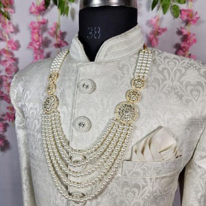 May include: A cream-colored, patterned jacket with a pearl necklace. The necklace features multiple strands of pearls and ornate gold-colored circular accents. The jacket has decorative buttons and a pocket square.