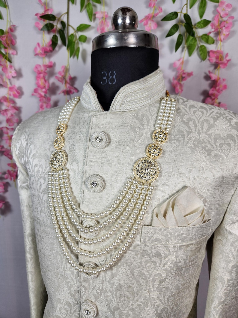 May include: A cream-colored, patterned jacket with a pearl necklace. The necklace features multiple strands of pearls and ornate gold-colored circular accents. The jacket has decorative buttons and a pocket square.