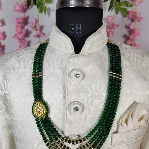 Puede incluir: Collar de cuentas verdes de varias hebras con detalles dorados y un colgante decorativo. El collar se muestra en un maniqu&iacute; que lleva una chaqueta blanca estampada con un pa&ntilde;uelo de bolsillo. El collar es una joya llamativa.