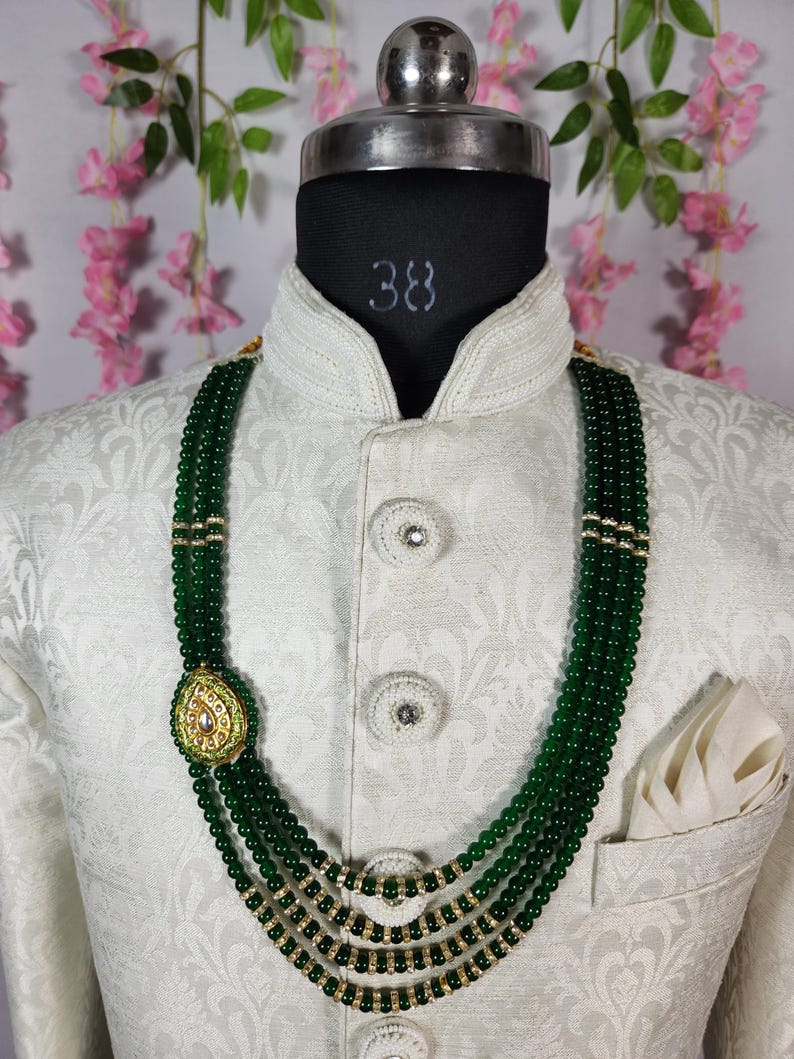 Puede incluir: Collar de cuentas verdes de varias hebras con detalles dorados y un colgante decorativo. El collar se muestra en un maniqu&iacute; que lleva una chaqueta blanca estampada con un pa&ntilde;uelo de bolsillo. El collar es una joya llamativa.