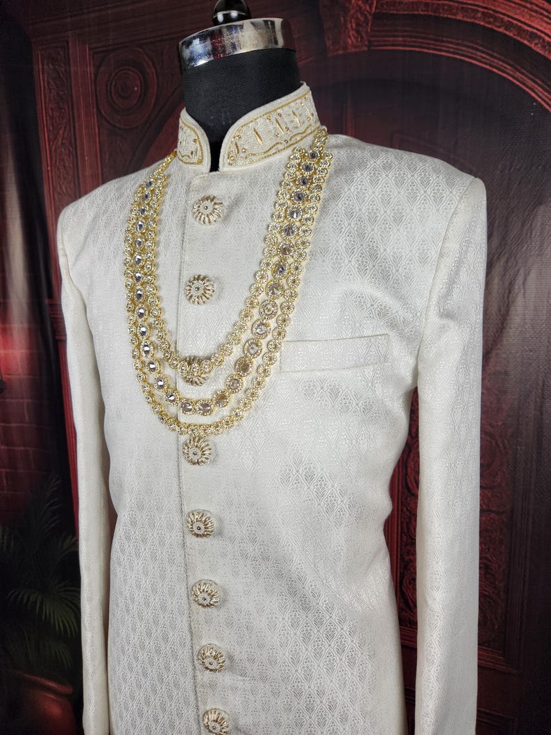 K&ouml;nnte beinhalten: Cremefarbene formelle Jacke mit aufwendiger Goldstickerei an Kragen und Kn&ouml;pfen. Eine mehrreihige goldene Halskette mit klaren Edelsteinen drapiert die Jacke. Die Jacke hat ein subtiles, strukturiertes Muster.
