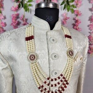 K&ouml;nnte beinhalten: Cremefarbene Perlenkette mit rubinroten Akzenten und dekorativen Anh&auml;ngern, pr&auml;sentiert auf einer wei&szlig;en Jacke. Die Kette hat vier Str&auml;nge und ist ein elegantes Accessoire f&uuml;r formelle Anl&auml;sse.