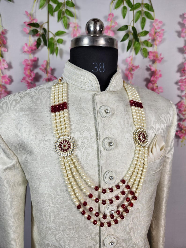 K&ouml;nnte beinhalten: Cremefarbene Perlenkette mit rubinroten Akzenten und dekorativen Anh&auml;ngern, pr&auml;sentiert auf einer wei&szlig;en Jacke. Die Kette hat vier Str&auml;nge und ist ein elegantes Accessoire f&uuml;r formelle Anl&auml;sse.