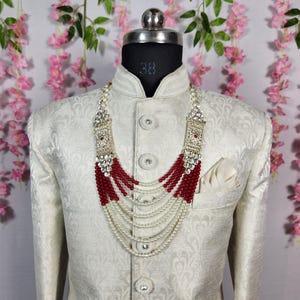 May include: A cream-colored jacket with a detailed pattern, paired with a multi-strand necklace. The necklace features red beads, white pearls, and ornate silver-toned accents. A white pocket square is visible in the jacket pocket.