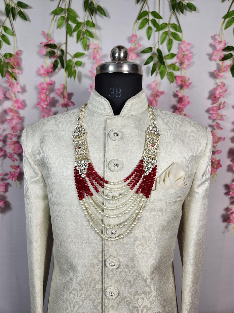 May include: A cream-colored jacket with a detailed pattern, paired with a multi-strand necklace. The necklace features red beads, white pearls, and ornate silver-toned accents. A white pocket square is visible in the jacket pocket.