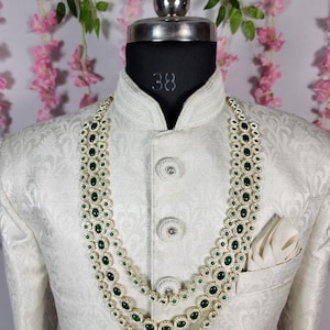Pode incluir: Um colar de tr&ecirc;s fios com pedras verde esmeralda e um casaco branco ornamentado. O colar apresenta pedras verdes ovais engastadas em metal dourado, rodeadas por pedras brancas menores. O casaco tem gola alta e um bolso com um len&ccedil;o branco.