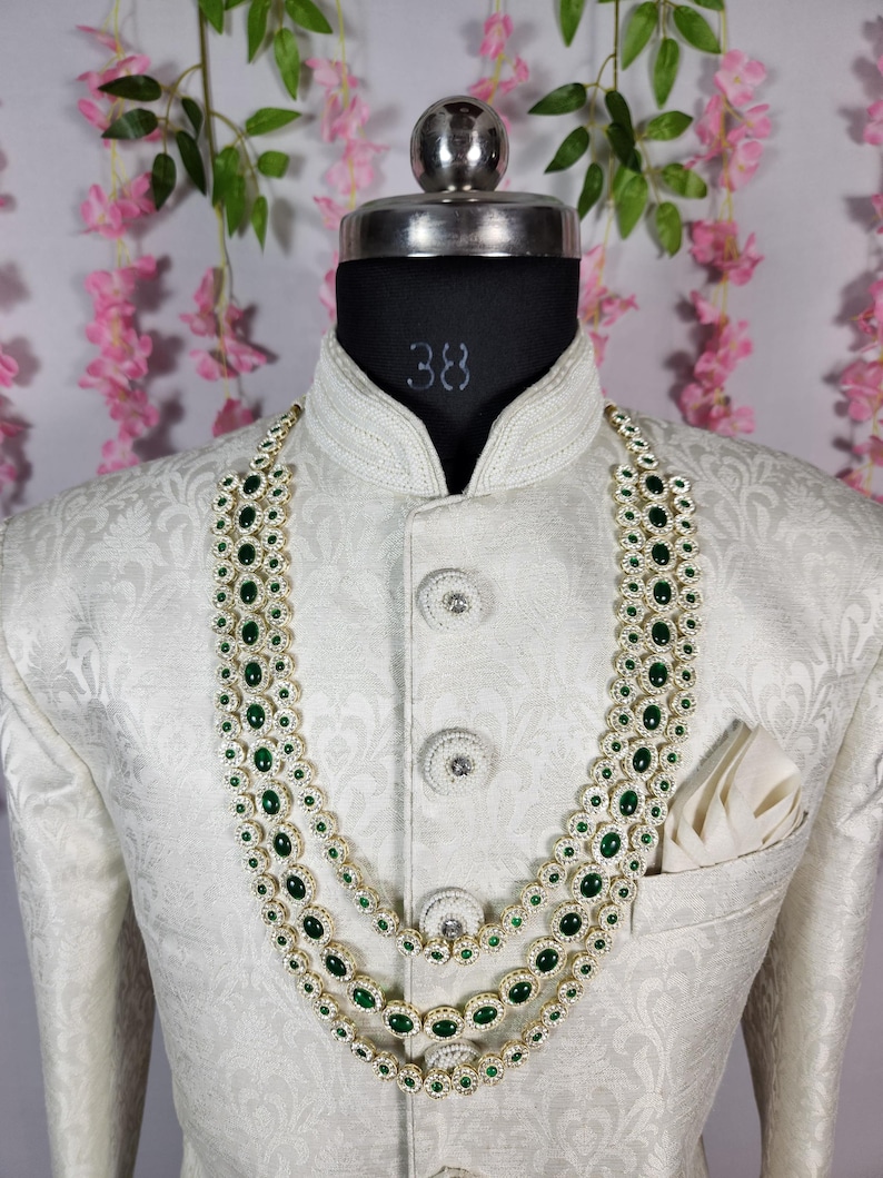 Pode incluir: Um colar de tr&ecirc;s fios com pedras verde esmeralda e um casaco branco ornamentado. O colar apresenta pedras verdes ovais engastadas em metal dourado, rodeadas por pedras brancas menores. O casaco tem gola alta e um bolso com um len&ccedil;o branco.