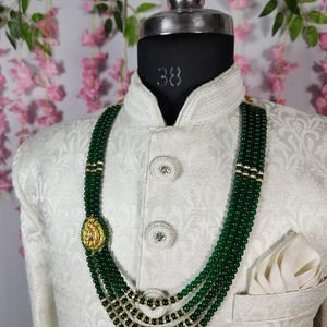 Puede incluir: Un collar de cuentas verdes con detalles dorados y un colgante decorativo se muestra en un maniqu&iacute;. El collar es en capas y se usa sobre una chaqueta blanca con un pa&ntilde;uelo de bolsillo. El fondo presenta flores rosas.
