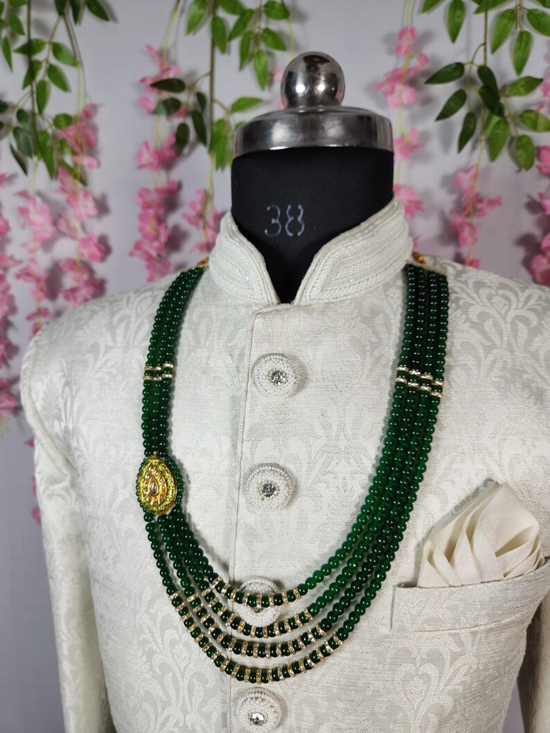 Puede incluir: Un collar de cuentas verdes con detalles dorados y un colgante decorativo se muestra en un maniqu&iacute;. El collar es en capas y se usa sobre una chaqueta blanca con un pa&ntilde;uelo de bolsillo. El fondo presenta flores rosas.