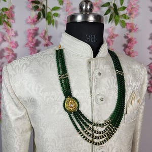 Puede incluir: Un collar de cuentas verde oscuro con un colgante dorado se muestra en un maniqu&iacute; con una chaqueta color crema. El collar presenta m&uacute;ltiples hilos y un colgante dorado con una piedra central. La chaqueta tiene un sutil estampado floral.