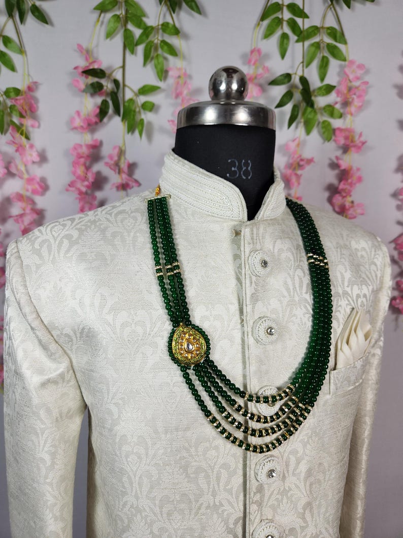 Puede incluir: Un collar de cuentas verde oscuro con un colgante dorado se muestra en un maniqu&iacute; con una chaqueta color crema. El collar presenta m&uacute;ltiples hilos y un colgante dorado con una piedra central. La chaqueta tiene un sutil estampado floral.