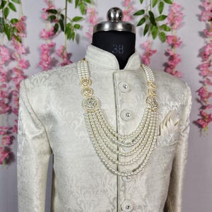 May include: A cream-colored, patterned jacket with a matching pocket square and a multi-strand pearl necklace with gold-toned accents. The jacket is buttoned and displayed on a mannequin.