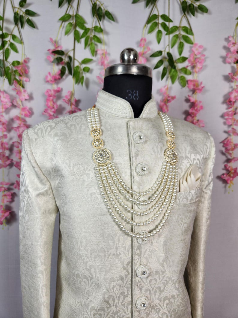 May include: A cream-colored, patterned jacket with a matching pocket square and a multi-strand pearl necklace with gold-toned accents. The jacket is buttoned and displayed on a mannequin.