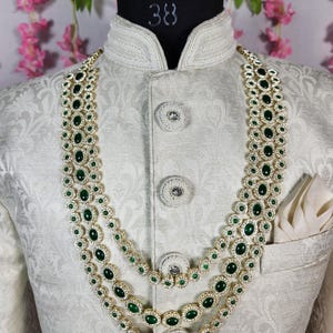 Pode incluir: Um colar de tr&ecirc;s fios com pedras preciosas ovais verde esmeralda e pequenas pedras transparentes engastadas em metal dourado. O colar &eacute; exibido em um casaco branco com estampa floral e um len&ccedil;o de bolso.