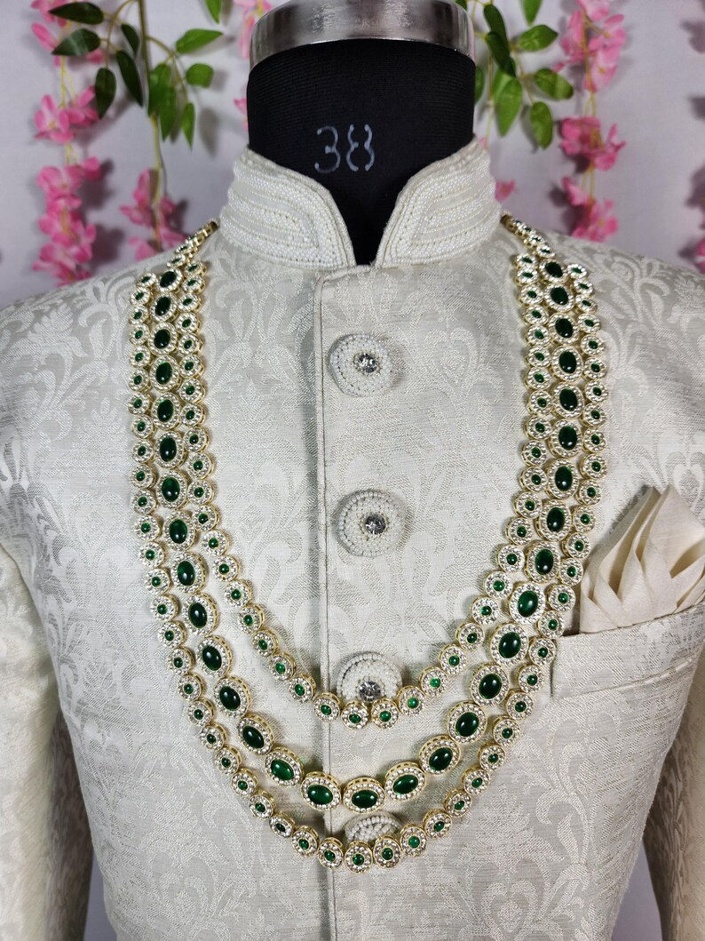 Pode incluir: Um colar de tr&ecirc;s fios com pedras preciosas ovais verde esmeralda e pequenas pedras transparentes engastadas em metal dourado. O colar &eacute; exibido em um casaco branco com estampa floral e um len&ccedil;o de bolso.