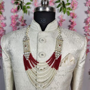 May include: A cream-colored jacket with a floral pattern, worn by a mannequin. A layered necklace with red beads and white pearls is displayed. The necklace has a silver-colored rectangular top with clear stones.