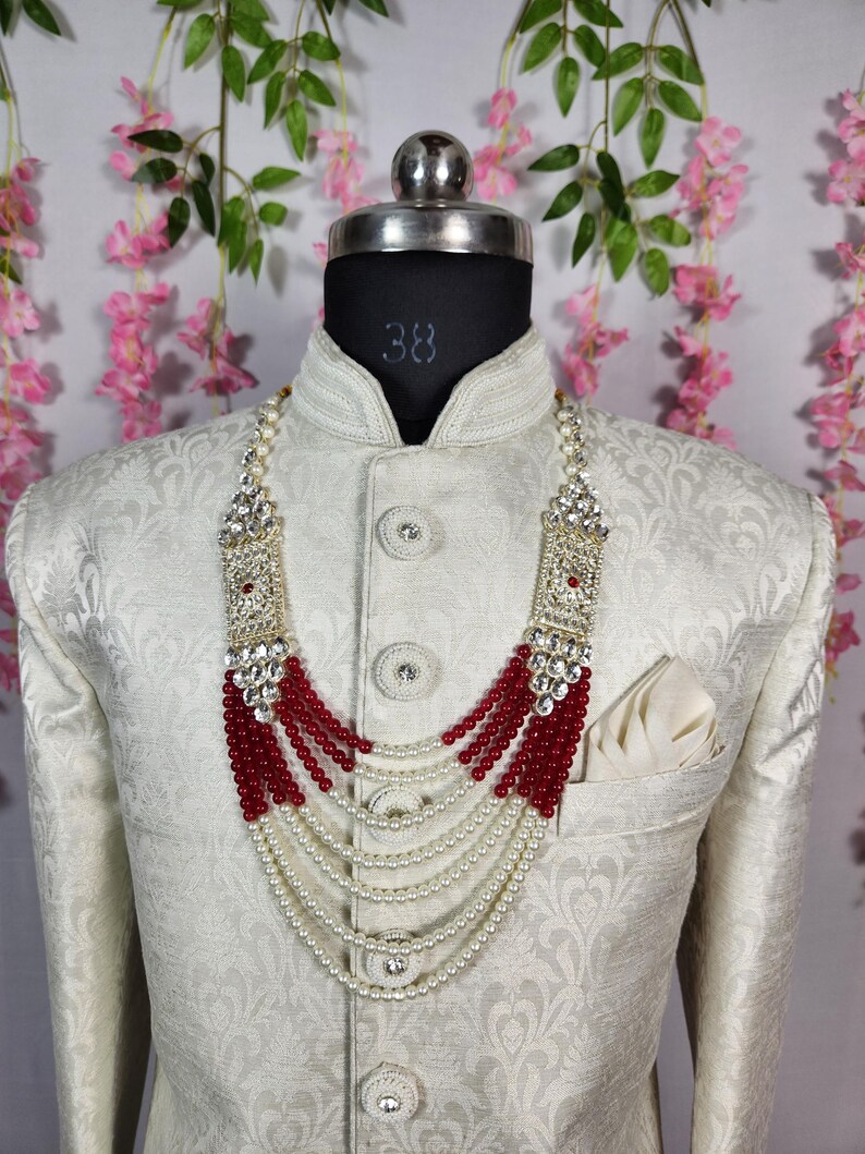 May include: A cream-colored jacket with a floral pattern, worn by a mannequin. A layered necklace with red beads and white pearls is displayed. The necklace has a silver-colored rectangular top with clear stones.