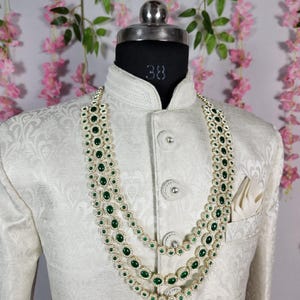 Pode incluir: Um colar de tr&ecirc;s fios com pedras preciosas verde esmeralda e um casaco branco. O colar &eacute; em camadas, com cada fio apresentando uma s&eacute;rie de pedras verdes ovais. O casaco &eacute; branco com um padr&atilde;o floral sutil e bot&otilde;es decorativos.