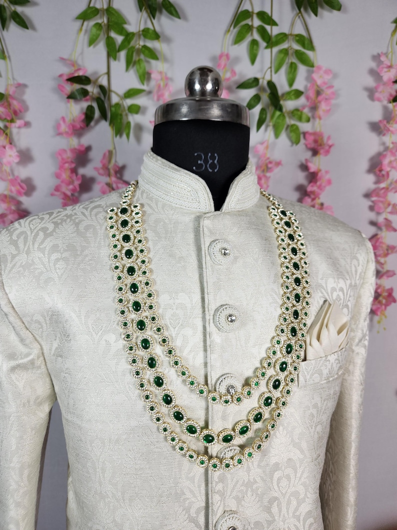 Pode incluir: Um colar de tr&ecirc;s fios com pedras preciosas verde esmeralda e um casaco branco. O colar &eacute; em camadas, com cada fio apresentando uma s&eacute;rie de pedras verdes ovais. O casaco &eacute; branco com um padr&atilde;o floral sutil e bot&otilde;es decorativos.