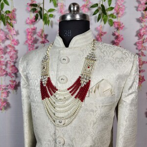 May include: A cream-colored, patterned jacket with a mandarin collar and a pocket square. A multi-strand necklace with white beads and red accents is draped over the jacket. The necklace has ornate silver-colored embellishments.