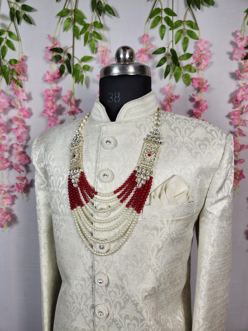 May include: A cream-colored, patterned jacket with a mandarin collar and a pocket square. A multi-strand necklace with white beads and red accents is draped over the jacket. The necklace has ornate silver-colored embellishments.