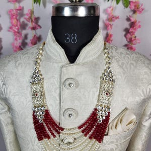 May include: A cream-colored jacket with a detailed pattern is worn by a mannequin. A multi-strand necklace with white pearls, red beads, and silver accents is displayed. The mannequin's neck is marked with the number 38.
