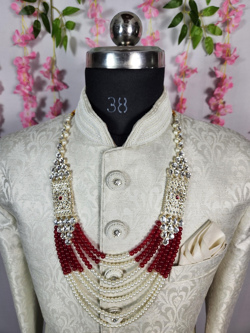 May include: A cream-colored jacket with a detailed pattern is worn by a mannequin. A multi-strand necklace with white pearls, red beads, and silver accents is displayed. The mannequin's neck is marked with the number 38.