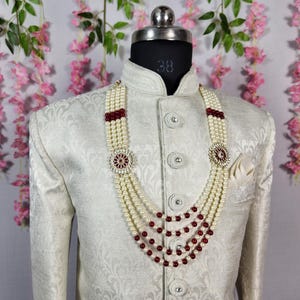 K&ouml;nnte beinhalten: Eine cremefarbene Jacke mit detailliertem Muster wird von einer Schaufensterpuppe getragen. Eine mehrstr&auml;ngige Perlenkette mit roten Akzenten und einem dekorativen Anh&auml;nger wird gezeigt. Ein wei&szlig;es Einstecktuch ist sichtbar.