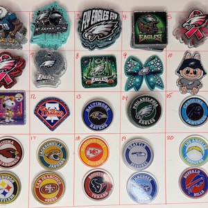 May include: A selection of glittery charms with various sports team logos, including the Philadelphia Eagles, Baltimore Ravens, and Kansas City Chiefs. The charms are round, square, and shaped, with vibrant colours and team emblems.