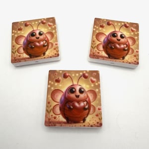 Op de afbeelding: Vier vierkante tegels met een cartoon lieveheersbeestje ontwerp. Het lieveheersbeestje is bruin met een hartvormig lichaam en houdt een hart vast. De achtergrond is lichtbruin met harten en bloemen.