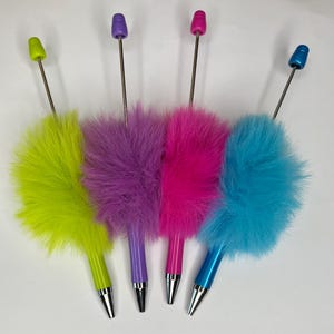 May include: Four colorful pens with fluffy feather accents. The pens have a variety of colors, including green, purple, pink, and blue. Each pen has a silver tip and a matching colored top.
