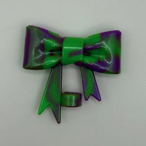 May include: A decorative bow with a vibrant color scheme. The bow features a mix of green and purple hues, creating a striking visual contrast. The bow's design includes a central knot and flowing tails, adding to its ornamental appeal.