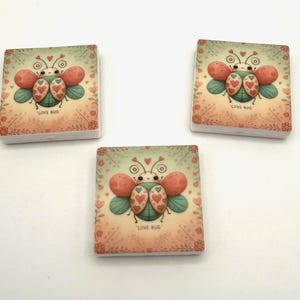 Op de afbeelding: Drie vierkante decoratieve tegels met een cartoon love bug-ontwerp. De kever heeft rode en groene vleugels, een hartvormig lichaam en een lachend gezicht. De tegels hebben een bloemenrand en de woorden "LOVE BUG".