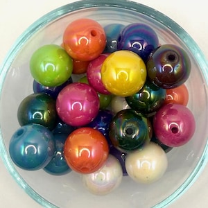 May include: A selection of iridescent, spherical beads in a clear glass bowl. The beads are in various colours, including orange, pink, green, yellow, and blue. Each bead has a hole through the centre. The beads have a glossy, reflective surface.