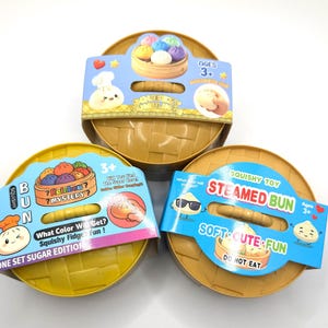May include: Three round, yellow, woven-textured plastic containers, each featuring a colorful cardboard sleeve. The sleeves display cartoon steamed buns, the text Squeezy Dumpling, Squishy Bun, and Steamed Bun, along with the age rating 3+.