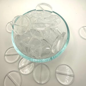 May include: A clear glass bowl filled with transparent, circular, pill-shaped objects. Each object has a line bisecting it. Some objects are scattered outside the bowl on a white surface.