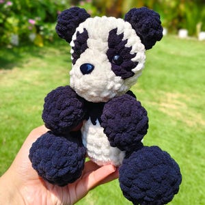 Pode incluir: Um panda de pelúcia feito à mão, com um corpo branco e detalhes em azul escuro. O panda tem orelhas, patas e marcas oculares em azul escuro. É feito de fio macio e texturizado e tem olhos de botão azuis.