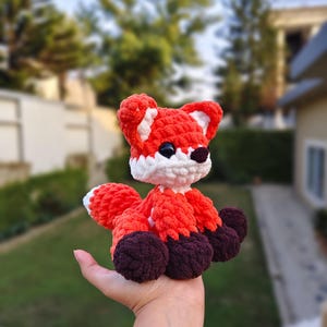 May include: A handmade crochet fox toy in shades of orange, white, and brown. The fox has large black eyes and a fluffy tail. The toy is held in a hand, showcasing its size and detail. The fox is a cute and cuddly stuffed animal.