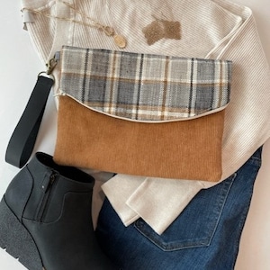 May include: A brown corduroy and plaid clutch with a zipper closure and a black strap. The clutch is laying on a white sweater, blue jeans, and black ankle boots.