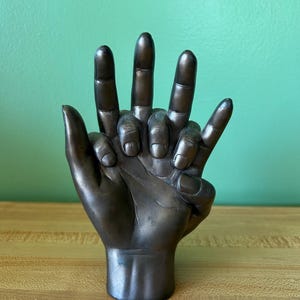 Puede incluir: Escultura de color bronce de una mano con los dedos extendidos. La escultura es una representación detallada de una mano humana, con nudillos y detalles de uñas visibles. La mano está posada en un gesto de apoyo u ofrenda.